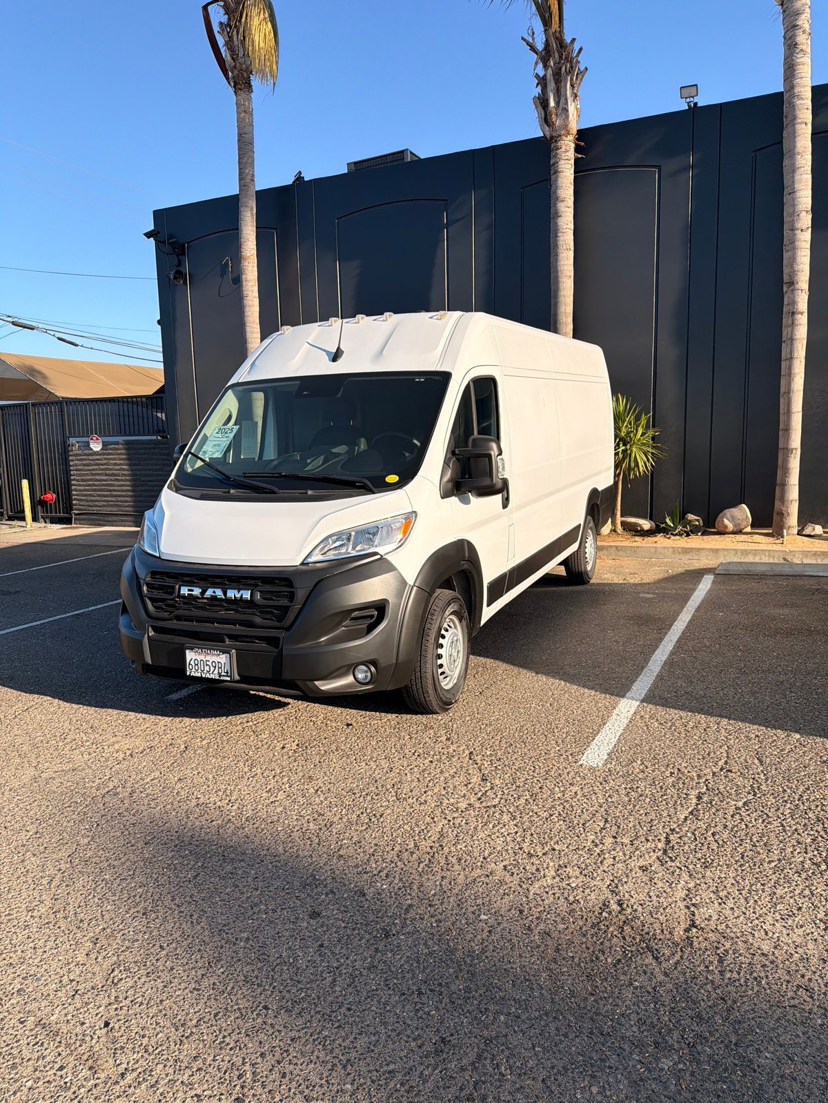 White RAM ProMaster van parked in a lot with black warehouse building and palm trees in background
